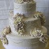 The WC-022 is a three-tiered white wedding cake with intricate piping, lace details, and cream and pale pink sugar roses, presented on a gold cake board against a light wood background.