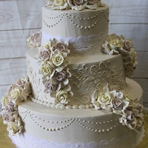 The WC-022 is a three-tiered white wedding cake with intricate piping, lace details, and cream and pale pink sugar roses, presented on a gold cake board against a light wood background.