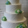 Product WC-011: A four-tier white cake with diagonal textured frosting, adorned with blue, white, and green buttercream roses and leaves, presented on a silver cake board against a wooden background.