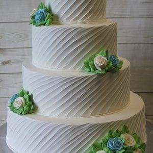 Product WC-011: A four-tier white cake with diagonal textured frosting, adorned with blue, white, and green buttercream roses and leaves, presented on a silver cake board against a wooden background.