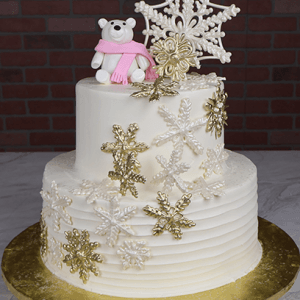 The TC-000A is a two-tiered white cake with gold and white snowflakes, topped with a fondant polar bear in a pink scarf, large snowflake decorations, and presented on a gold cake board.