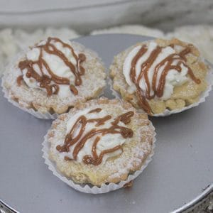 A slice of apple caramel cheesecake topped with powdered sugar and drizzled caramel, served on a round silver platter.
