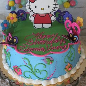 The SC-026 Hello Kitty cake features colorful flowers, butterflies, and a Hello Kitty topper. “Happy Birthday Zinnia Vuong” is written in pink icing. Multicolored balloon decorations appear in the background.