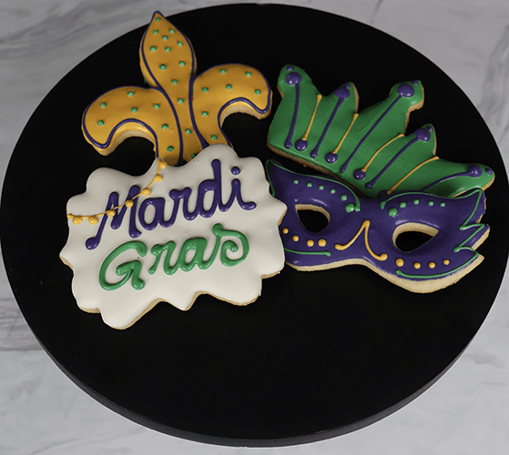 CE-007 Mardi Gras features three decorated cookies on a black plate: a yellow and green fleur-de-lis, a green and purple mask, and a white cookie with “Mardi Gras” in purple and green icing.
