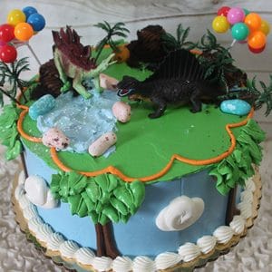 The SC-065 Dinosaur cake is round and blue, topped with trees, clouds, green frosting, two dinosaur figurines, candy eggs, balloon toppers, a small pond, and plastic plants for a fun prehistoric theme.