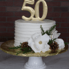 The BD-132 is a white frosted cake on a pedestal stand with a gold "50" topper, white fondant flowers, pinecones, and green foliage, set against a brick wall background.