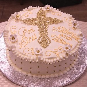 A round white cake (RC-019) decorated with gold accents, a large ornate gold cross on top, "God Bless" and "Katherine Anna Tatiana" in gold icing, and adorned with small white and gold flowers.