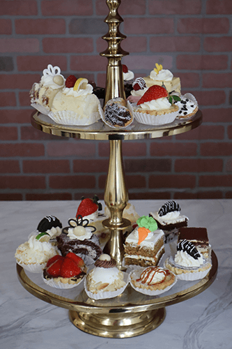 The AP-007 Assorted Mini Pastries Chef's Choice are elegantly arranged on a two-tier gold stand, featuring mini cakes topped with strawberries, chocolate, and cream, set against a brick wall and marble surface.
