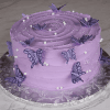 The BD-060I features a round cake with smooth light purple frosting, adorned with purple butterfly toppers, white sugar pearls, and a swirl pattern on top, presented on a silver floral-patterned cake board.