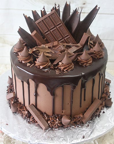 The BD-026A Chocolate Lovers Dream features a rich chocolate cake with drip, topped with bars, shards, swirls, and shavings, plus extra chocolate pieces around the base—all elegantly displayed on a silver board.