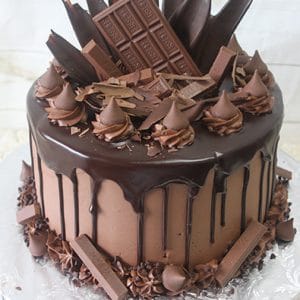 The HW-014 Chocolate Lovers Dream features a round chocolate cake with chocolate frosting, dark ganache drips, and a topping of assorted chocolate bars, shards, swirls, and curls on a silver cake board.