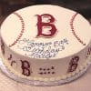 The ST-003 Red Sox cake is round, decorated like a baseball with red stitching and a large red B on top. “Happy 50th Birthday Matty” is written in blue icing, with baseballs and red B’s on the sides.