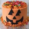 The HW-017 Trick or Treat is a round jack-o’-lantern cake with orange icing, black fondant facial features, and a topping of colorful gummy candies and sprinkles. Halloween sprinkles and gummy worms decorate the base.