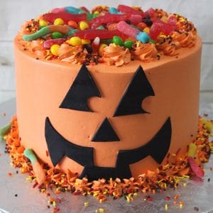 The HW-017 Trick or Treat is a round jack-o’-lantern cake with orange icing, black fondant facial features, and a topping of colorful gummy candies and sprinkles. Halloween sprinkles and gummy worms decorate the base.