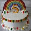 The CB-022C is a white frosted cake with colorful gummy bears, rainbow sprinkles, a large yellow number 5 topper, a paper rainbow backdrop, and sits on a silver board.