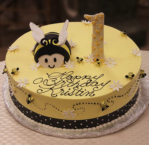 The CB-012J is a round yellow birthday cake with small bees, white flowers, a large bee face and wings, plus a number “1” topper. “Happy Birthday Kristin” is written in cursive icing.