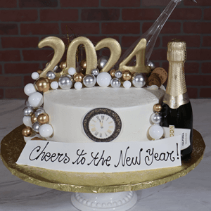 The NY-001A cake features “2024” numbers, gold, silver, and white spheres, a champagne bottle, a clock, and a “Cheers to the New Year!” sign on a gold stand against a brick background.