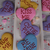 CE-020I Valentine's features heart-shaped cookies in pastel colors with messages like “KISS ME,” “CUTIE PIE,” and “BE MINE” on a white tray, surrounded by more decorated cookies and small candy hearts.