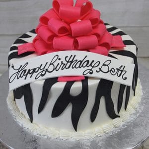 The BD-037A Zebra Bow Pink is a round cake with black zebra stripes, topped with a large pink bow and a white "Happy Birthday Beth" banner in cursive. It is presented on a silver platter.