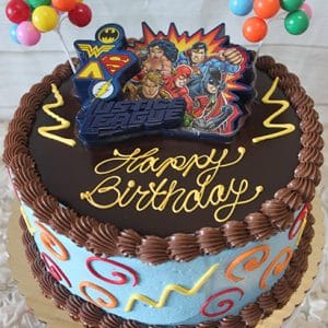 The SC-075 Justice League is a round birthday cake with blue icing, colorful swirls, chocolate frosting, “Happy Birthday” in yellow, Justice League superhero toppers, and candy balloon decorations.