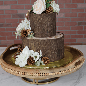 The WD-001D is a two-tier rustic cake with tree bark design, decorated with white and blush artificial flowers, pine cones, and gold accents, presented on a round wooden tray against a brick wall backdrop.