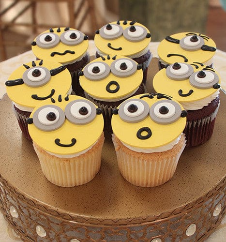 Eight CC-022 Chocolate cupcakes with white chocolate mousse filling, topped with yellow Minion-themed fondant, each featuring unique expressions and large candy eyes, arranged on a round cake stand.