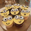 Nine CC-023 Gold cupcakes with chocolate filling, decorated with yellow icing and Minion cartoon faces, each showing a unique expression, arranged on a brown plate.
