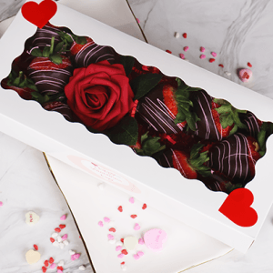 The VC-000A One dozen chocolate covered strawberries are presented in a white box with a clear window and red heart decorations, accompanied by a red artificial rose. The box sits on marble with scattered candy hearts and sprinkles.