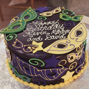 The BD-165 is a round cake with green, yellow, and purple masquerade masks and stars, finished with white icing that reads “Happy Birthday Kevin, Martyn, and David.”.