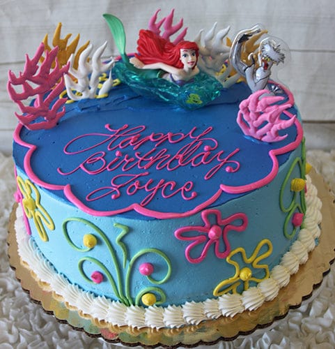 The SC-016A Little Mermaid cake is blue, decorated with colorful seaweed, shells, flowers, and topped with an Ariel figure. “Happy Birthday Joyce” is written in pink icing.