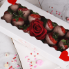 A white box with a clear window holds the VC-000B One Dozen Milk Chocolate Covered Strawberries and a red artificial rose, decorated with red hearts. The box is on a plate surrounded by heart-shaped sprinkles and candy hearts.