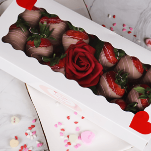 A white box with a clear window holds the VC-000B One Dozen Milk Chocolate Covered Strawberries and a red artificial rose, decorated with red hearts. The box is on a plate surrounded by heart-shaped sprinkles and candy hearts.