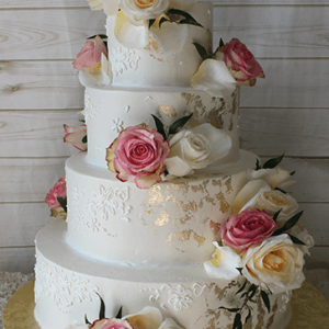 The WD-004 is a four-tiered white wedding cake with gold leaf accents, topped with pink, white, and yellow roses. It’s presented on a gold cake board against a wooden background.