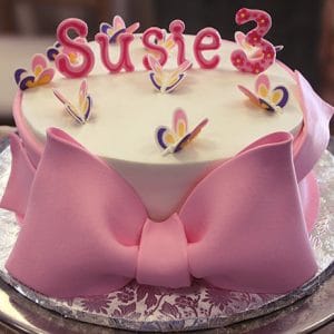 The CB-024A is a round cake with a large pink fondant bow, butterfly toppers, "Susie" in pink letters, and a pink number 3 candle—perfect for celebrating a third birthday.