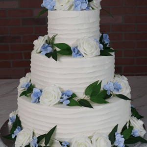 The WD-001H is a three-tiered cake with white frosting, adorned with white roses, blue flowers, and green leaves, displayed against a brick wall background.