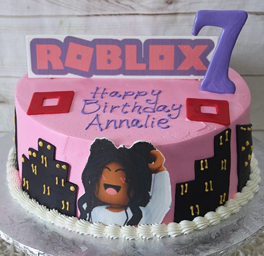 CB-011B: A pink Roblox-themed birthday cake with cityscape décor, a character image, “Happy Birthday Annalie” message, topped with a purple number 7 and the Roblox logo.