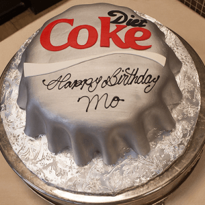 A round cake designed as a silver Diet Coke bottle cap, featuring "Happy Birthday Mo" in black icing. The BD-025 Diet Coke cake sits on a platter with a silver pattern.