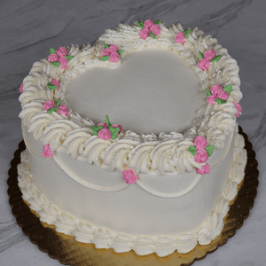 The BD-023D Heart shape one size cake features white frosting, piped white borders, and pink and green floral accents, presented on a gold cake board atop a marble surface.