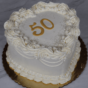 A heart-shaped white cake (BD-023C) with ornate white frosting, silver sprinkles, and a gold "50" topper on a gold board, set against a marble background.