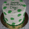 The STP-002 St. Patrick's Day 6" Cake features green shamrocks, green sprinkles, and "Happy St. Patricks Day!" written in green icing on top, all presented on a gold cake board. Available in one size only.