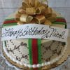 The BD-031 Designer Gold Bow is a round cake adorned with gold, green, and red ribbons, topped with a large gold bow and a white banner that reads "Happy Birthday Nicol" in elegant script.