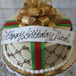 The BD-031 Designer Gold Bow is a round cake adorned with gold, green, and red ribbons, topped with a large gold bow and a white banner that reads "Happy Birthday Nicol" in elegant script.