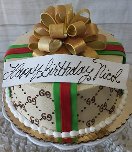 The BD-031 Designer Gold Bow is a round cake adorned with gold, green, and red ribbons, topped with a large gold bow and a white banner that reads "Happy Birthday Nicol" in elegant script.