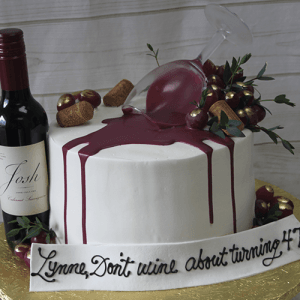 The BD-024R Wine Cabernet is a white cake decorated with a wine bottle, grapes, corks, and an overturned wine glass with red icing spilling out. "Lynne, Don't wine about turning 47!" is written on a fondant banner.