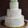 The WC-000A is a three-tiered white cake with floral designs and green leaves, topped with white roses, and displayed on a wooden tray against a brick wall background.