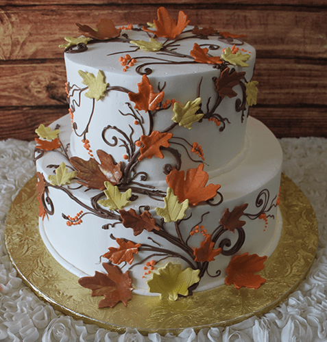 The TC-009 is a two-tiered white cake adorned with orange, yellow, and brown autumn leaves and vines, set on a gold board with a rustic wood background.