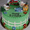 The SC-001 Farm Animals cake is round and green, decorated with a white fence, colorful flowers, cow, sheep, horse figurines, a farm scene topper, and "Happy Birthday" written on top in black icing.