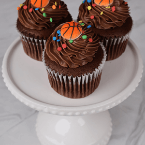 Three CC-097A chocolate cupcakes with white filling, topped with colorful sprinkles and small basketball decorations, are displayed on a white cake stand.