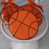 Three CE-002B Basketball-Shaped Cookies with orange icing and black lines are displayed on a white cake stand; two are wrapped in clear bags tied with orange bows.