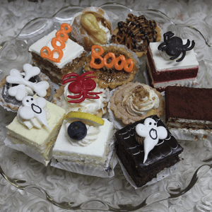A glass plate displays HW-019: assorted Halloween mini cakes and pastries topped with fondant spiders, ghosts, bones, and "Booo" in orange icing, all arranged on a textured white surface.